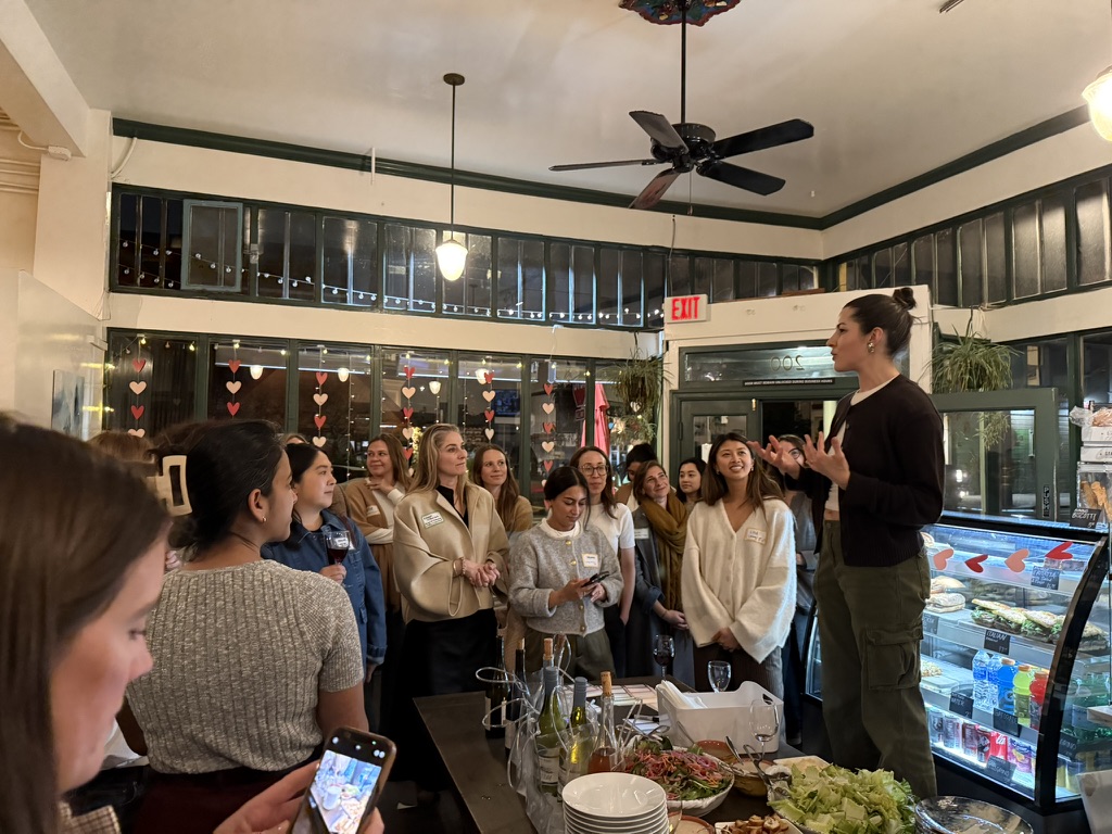 A group of women gathered at a Women + AI dinner, listening to a speaker next to a table of food.