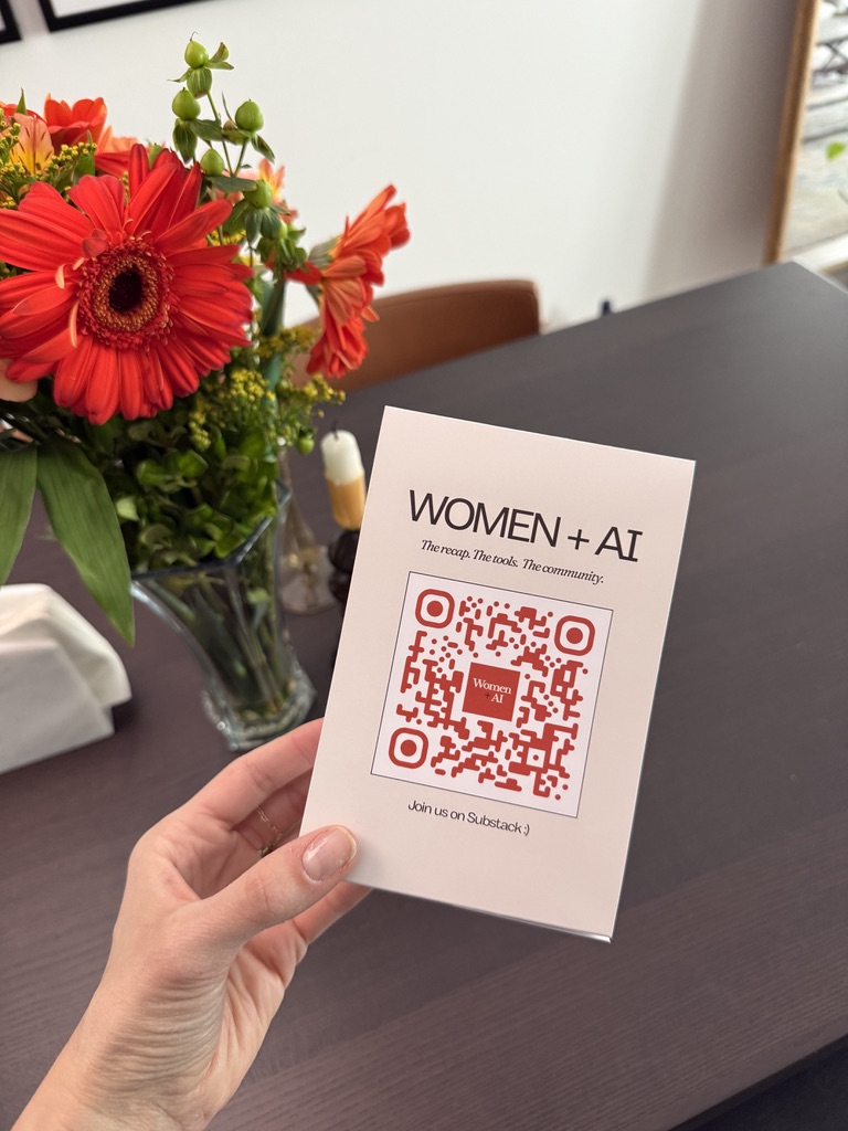 A hand holding a Women + AI Substack QR flyer next to red flowers.